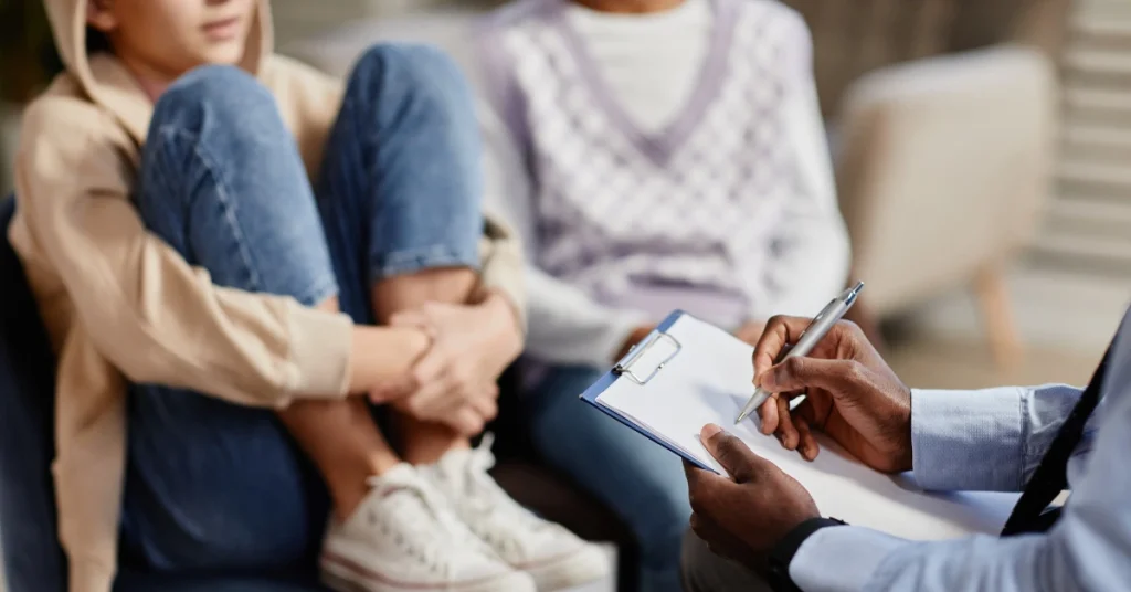 A therapist takes notes during a counseling session with two teens, one sitting with knees up and arms wrapped around their legs, conveying a serious tone.