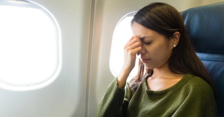 Woman sitting on an airplane with eyes closed and hand on forehead, showing signs of flight anxiety or stress while traveling.